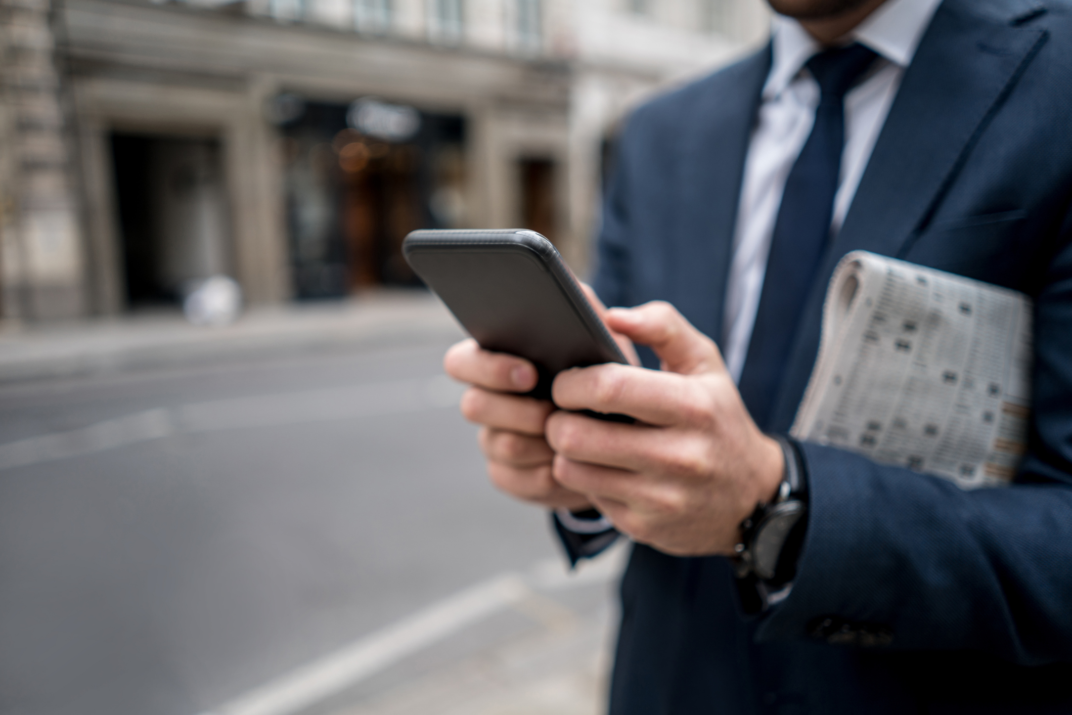 Close-up on a business man texting on the phone in the street and carrying the newspaper