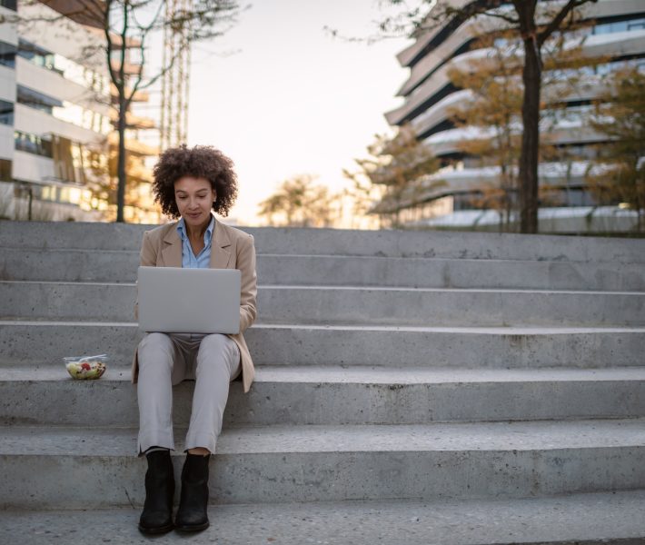 Beautiful mixed race young businesswoman sitting in a park on the bench during a lunch break, working on laptop and eating a vegetable salad.