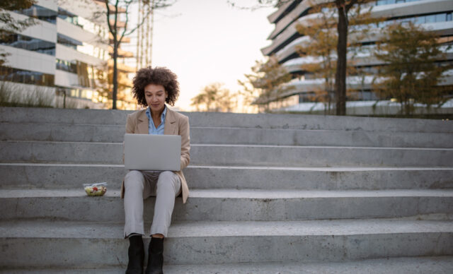 Beautiful mixed race young businesswoman sitting in a park on the bench during a lunch break, working on laptop and eating a vegetable salad.