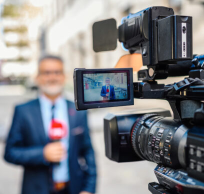 A male television reporter holding a microphone, is standing outdoors in front of a building. A male camera operator is filming him. Selective focus.
