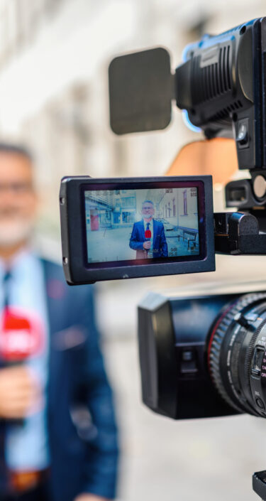 A male television reporter holding a microphone, is standing outdoors in front of a building. A male camera operator is filming him. Selective focus.