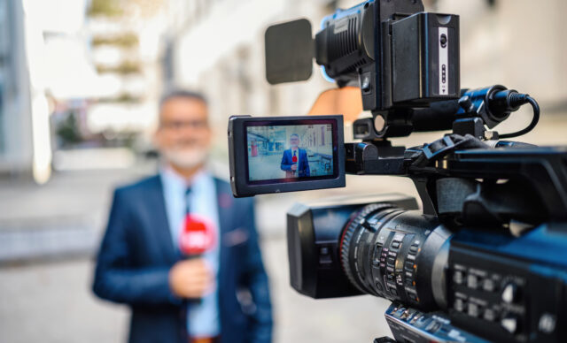 A male television reporter holding a microphone, is standing outdoors in front of a building. A male camera operator is filming him. Selective focus.