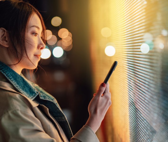 Young Asian woman using smartphone against illuminated digital display in city street at night. Blockchain technology. Trading cryptocurrency. NFT (Non-Fungible Token) investment.