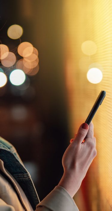 Young Asian woman using smartphone against illuminated digital display in city street at night. Blockchain technology. Trading cryptocurrency. NFT (Non-Fungible Token) investment.