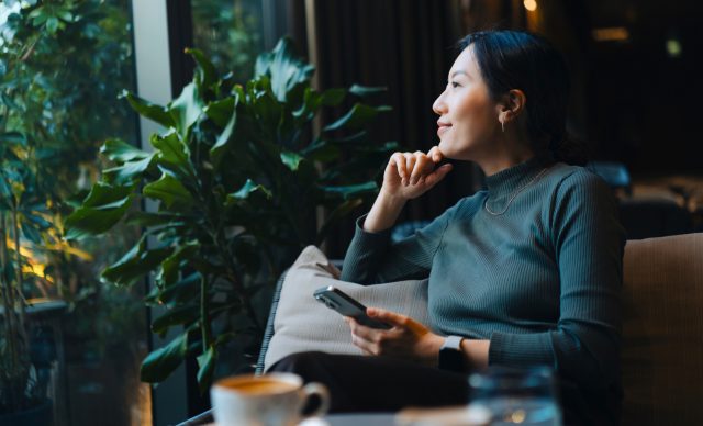 Confident young Asian woman with smartphone looking out through window while sitting in a cafe having coffee.