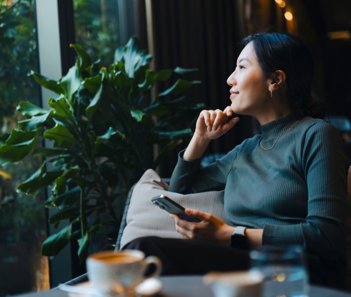 Confident young Asian woman with smartphone looking out through window while sitting in a cafe having coffee.