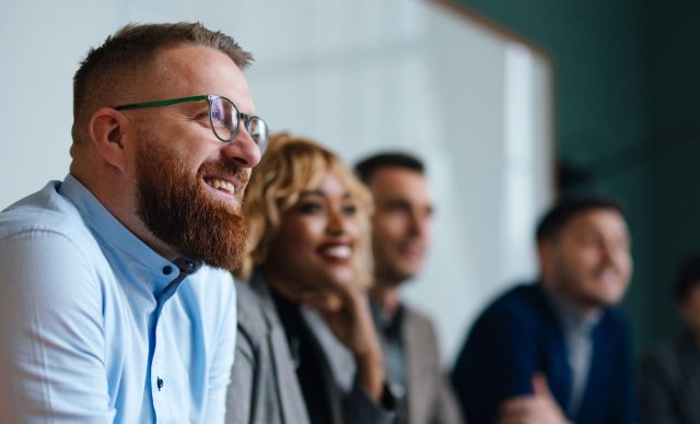 Smiling businessman and unrecognizable multi-ethnic employees sitting in a row at their company desk and listening to their boss.