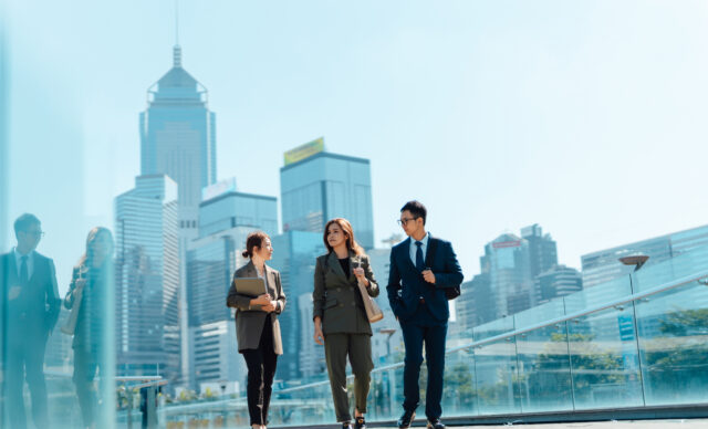 Three confident and successful Asian business people having a discussion on the go, walking in financial district against urban skyline. Teamwork and collaboration. Striving for success. Overcoming challenges and reaching goals.